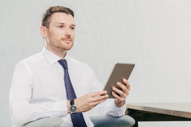 istock Thoughtful Caucasian male laywer thinks about something, holds modern tablet computer, wears white shirt with black tie, sits at his cabinet, waits for client. People, work, technology concept 1446949269