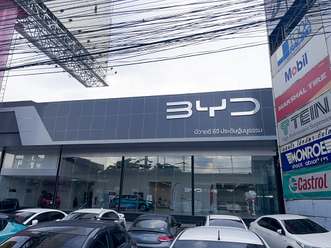 BYD car retail shop in Bangkok Ramintra. View from sidewalk over parking lot to store. Inside are some customers and a retail clerk staff