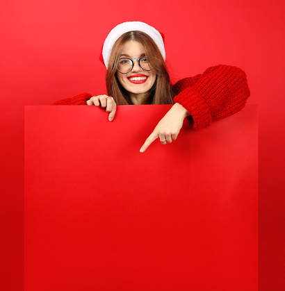 A cute smiling girl in a Santa hat and a Christmas sweater with a red advertising banner in her hands on a red background.