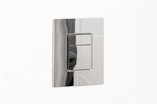 Metal chromed toilet flush button on white tile wall. Details in a bright modern bathroom