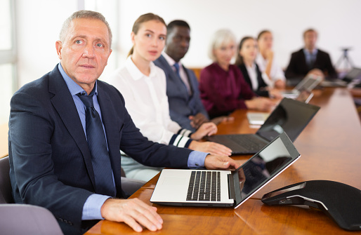 Mature senior white male manager attending business meeting in conference room and intently listening to colleague's report together with coworkers sitting in row behind him