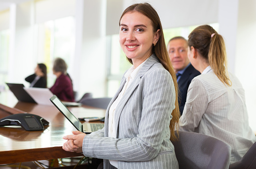 Positive young woman manager sitting at desk and looking back at camera during meeting with colleagues in conference room in company office.