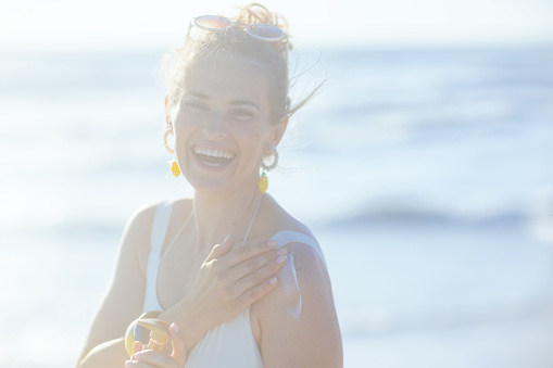 Portrait of happy elegant woman in white swimwear at the beach applying sunscreen.