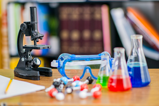 Various science supplies lay out on a students desk, including a microscope, beakers, flasks, protective eyewear, etc. in this concept still photo.