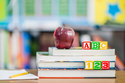 A stack of school books sit on a students desk with an apple on top and colorful blocks that read \