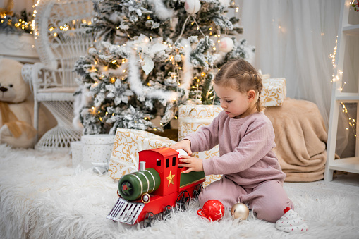 Little girl in pajamas playing with red retro train under the Christmas tree in home room. High quality photo