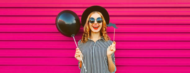 Portrait of funny cheerful young woman having fun holding black balloon, showing mustache on stick on pink background stock photo
