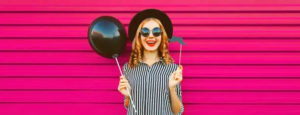 Portrait of funny cheerful young woman having fun holding black balloon, showing mustache on stick on pink background