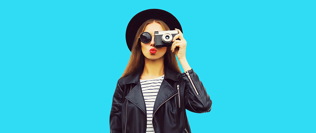 Stylish portrait of beautiful young woman photographer with film camera wearing black round hat, leather jacket isolated on blue background