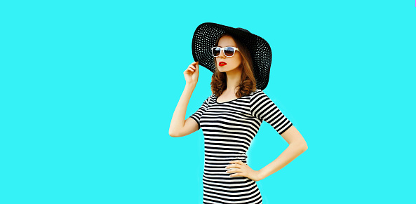 Portrait of beautiful woman model looking away wearing black round summer hat, striped dress isolated on blue background