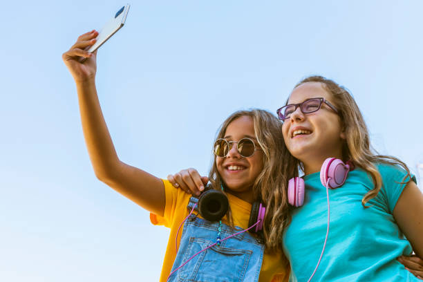 две кавказские молодые девушки в солнцезащитных очках и наушниках улыбаются и делают сел�фи со смартфоном на открытом воздухе. концепция об - teenager couple child blond hair стоковые фото и изображения