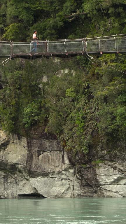 Young adult man crossing high suspension bridge above river.