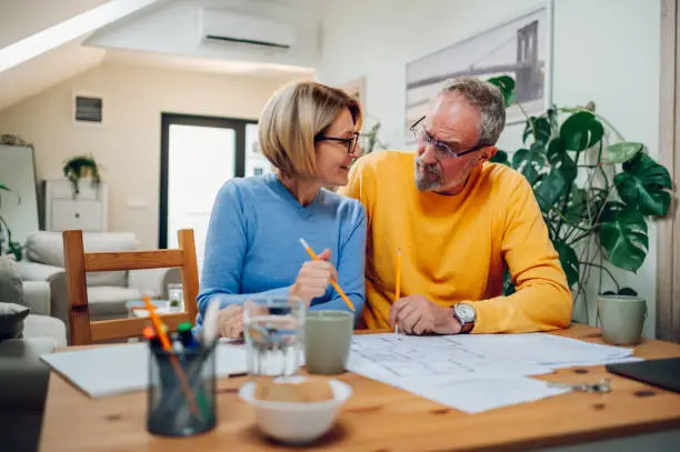 Senior married couple is actively discussing repairs in a new apartment or house while measuring the dimensions in the blueprints and sitting at the table and making notes.