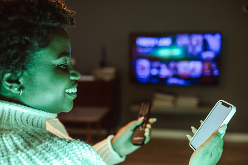 A cheerful smiling woman is watching TV and holding the remote control