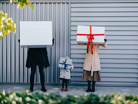 Anonymous shy father, mother and little daughter holding wrapped gift boxes while standing in a row in front of metal wall.
