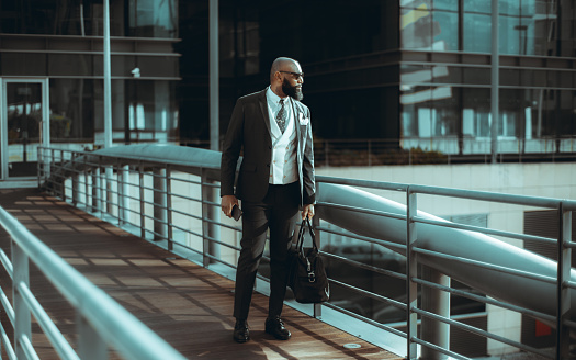 White-collar worker carrying a gym bag in one hand and a cell phone in the other. A bearded, bald black man wearing sunglasses, a dark suit, a tie and a white vest, exiting a contemporary building