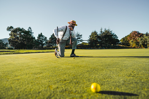 Bearded afro-descending man, all dressed up, wearing tailored grey pants, a white shirt, striped suspenders, with a hat. Kneeling on the floor of the golf course, leaning on his club, on a clear day