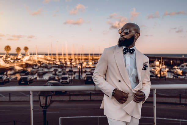 Stylish black male model at bay A stylish bearded black man poses at the golden hour; The African man is dressed in a beige suit with blue bow tie and white polka dots, and sunglasses, behind him the moored boats at bay rich man stock pictures, royalty-free photos & images