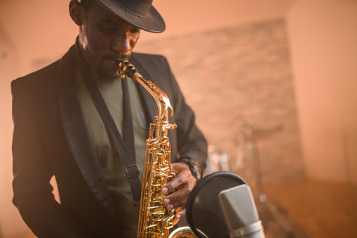 Black mid-adult male musician playing saxophone and recording songs in recording studio. Waist-up, looking away.