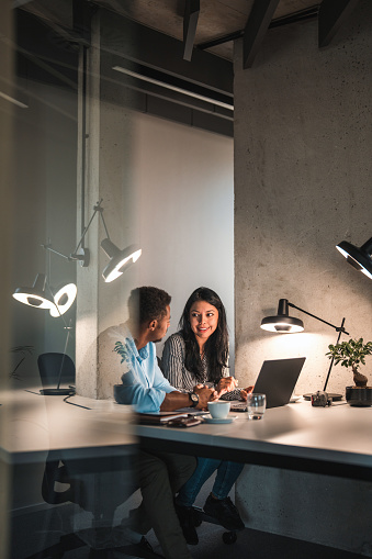 Good looking African American and Latin woman working hard and using laptop at work. They are looking face to face. The glass door is open reflecting the light from their desk lamp.