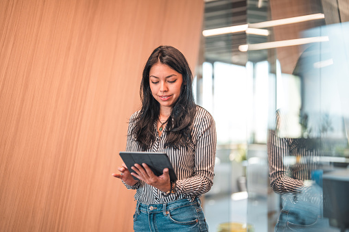 Front view portrait of attractive black hair businesswoman using a digital tablet. Female manager standing at her modern office and looking at her tablet.