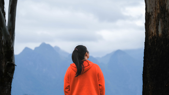Rear view of a young woman standing on the top of a calm mountain and rising hands on mountain in morning. Woman wearing a sweater enjoying the beauty of nature looking at the mountain in winter.