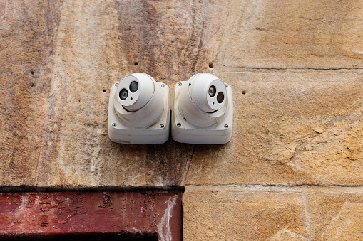 Two white security cameras on an old sandstone wall.