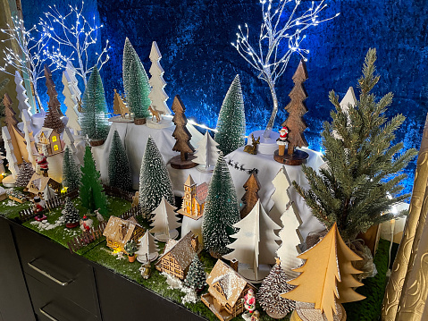 Stock photo showing a beautiful Christmas village display, which features numerous illuminated wooden houses. The houses have been placed on a fleece blanket to create a snowy mountain scene, in front of a blue velvet backdrop, complete with a forest of plastic and wooden trees.