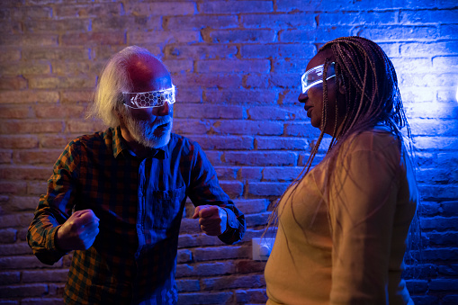 Senior multiracial couple looking at each other with virtual reality goggles in a neon light room. Focus on african american mature woman