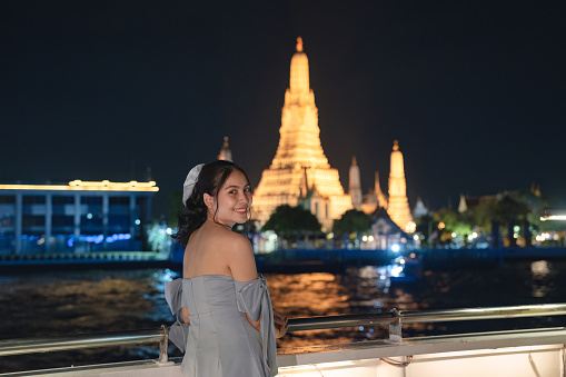 Beautiful young asian woman in dress standing and looking at camera on the deck with Wat Arun glowing in the night. Dinner cruise sailing on the Chao Phraya river in Bangkok