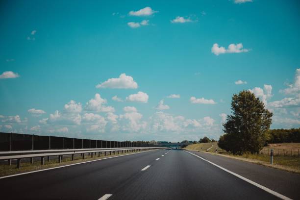 istock Empty asphalted road with white markings, railings, and trees beside it under blue sky with clouds 1447085417