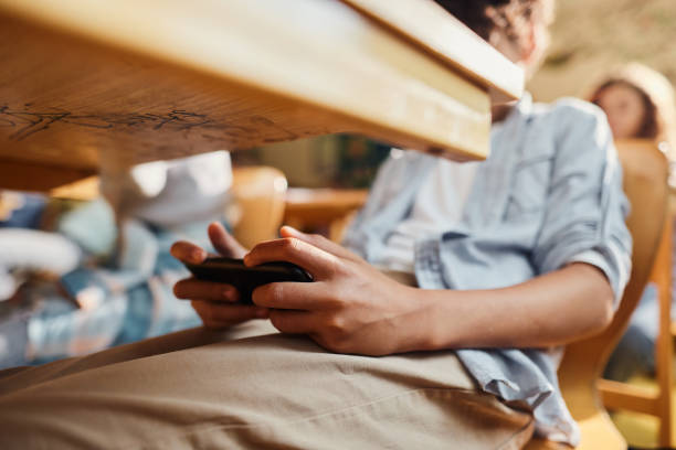 Secretly using cell phone on a class! Close up of unrecognizable black student using cell phone under the table during a class at elementary school. digital native stock pictures, royalty-free photos & images