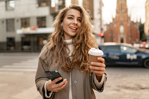 Young woman wearing autumn coat walking with smartphone and coffee cup in a city street, portrait
