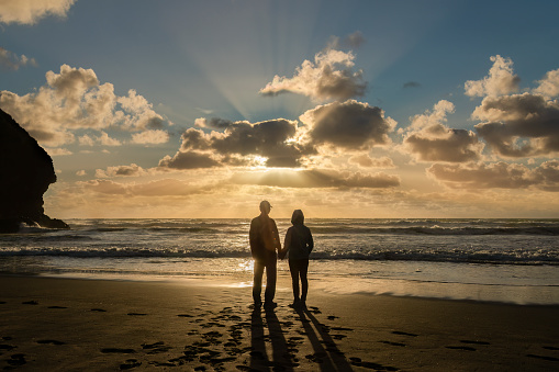 Couple watching sunset at Piha Beach, sunbeams shining through the clouds. Waitakere, Auckland