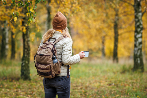 Woman with backpack and knit hat resting with hot drink during autumn hike in forest