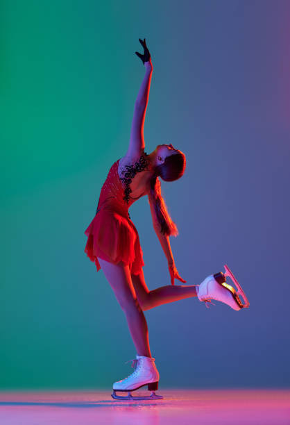 deportes de invierno. una patinadora artística femenina junior con traje de escenario rojo que muestra elementos de patinaje artístico base, movimientos aislados sobre fondo verde-azul degradado en luz de neón. - axel fotografías e imágenes de stock