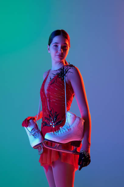 after workout. charming young teen girl in stage costume dress standing with skates isolated over gradient green-blue background in neon light. concept of skills, sport, hobbies - axel imagens e fotografias de stock