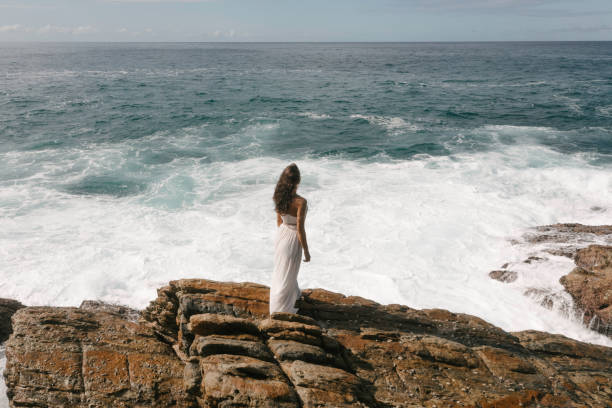 Aerial drone view of bride in an amazing dress standing on a cliff and looking at the sea Aerial drone view of beautiful bride in an amazing white dress standing on a cliff and looking at the sea. Epic scene with stormy waves and gorgeous woman model goddess of ocean stock pictures, royalty-free photos & images