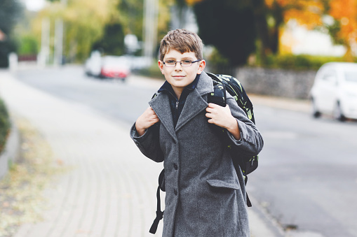 Happy kid boy with glasses and backpack or satchel. Schoolkid in stylish fashon coan on the way to middle or high school on cold autumn day. Healthy child outdoors on the street, on rainy day