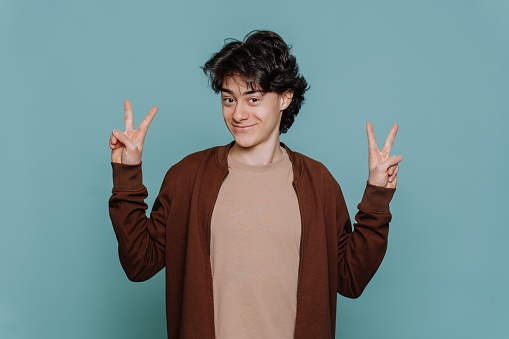Playful handsome boy in casual smiles looks at camera shows victory sign by both hands standing against turquoise studio backdrop. Brunette schoolboy passed exams. Happy teen winer.
