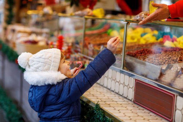 kleines süßes vorschulmädchen, das zuckerstangen von einem süßigkeitenstand auf dem weihnachtsmarkt kauft. glückliches kind auf traditionellem familienmarkt in deutschland. vorschulkind in bunter winterkleidung - romrodinka stock-fotos und bilder