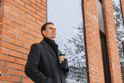 Low angle of businessman in coat with cup of takeaway coffee standing near building in winter city