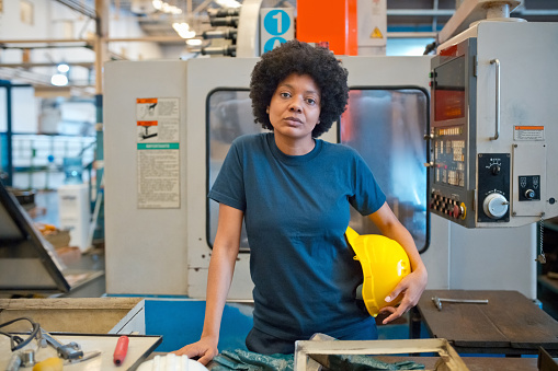 Portrait of female blue-collar worker standing in industry. Mature woman holding yellow work helmet looking at camera.
