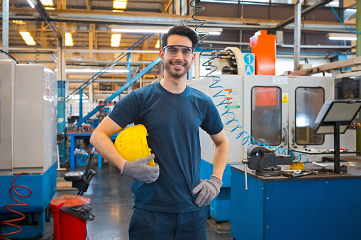 Portrait of smiling young male engineer wearing protective glasses and holding yellow helmet, standing with hand on hip in factory.