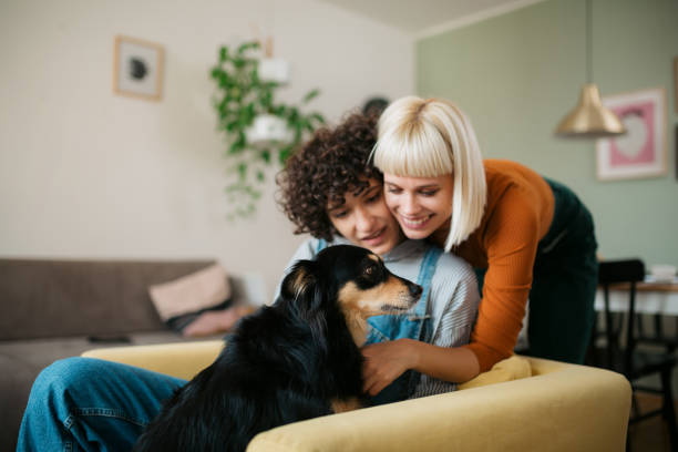 Happy couple of lovers relaxing at home with their pet dog Happy lesbian couple enjoying themselves at home, sitting in an armchair in their living room and snuggling with their pet dog, showing him affection lesbian couple embracing stock pictures, royalty-free photos & images
