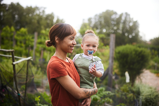 Smiling mother holding her cute little girl with a pacifier in her arms while standing outside in a community garden
