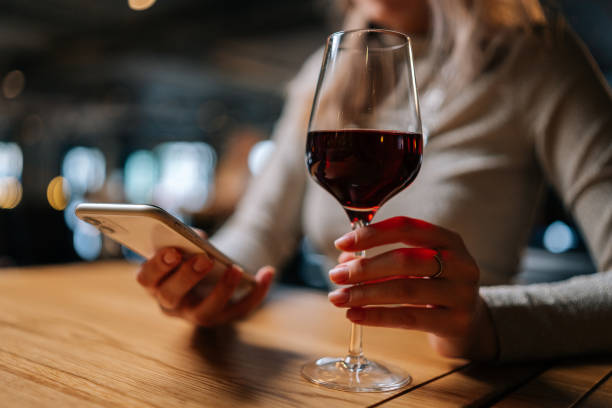 nahaufnahme einer nicht erkennbaren jungen frau, die ein smartphone benutzt und eine online-nachricht tippt, die am tisch sitzt und ein glas rotwein in der hand hält. - alcohol stock-fotos und bilder