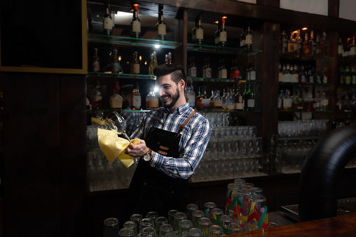 Young happy bartender smiling and polishing drinking glass in the cafeteria or bar with retro wooden look. Man cleaning glass with rug behind the counter.