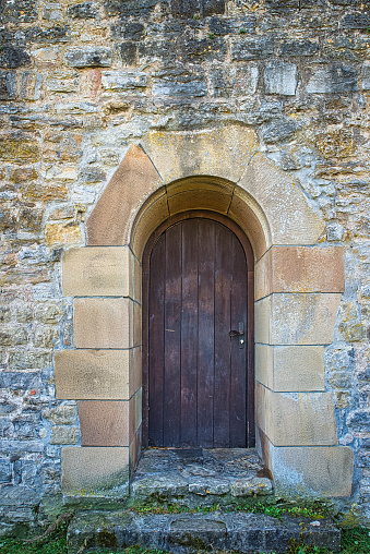 This wooden door forms the entrance to a medieval building. It is built of rough stones and the door frame was made of smooth sandstone blocks. In front of this light sandstone, the wooden door looks heavy and gloomy. The floor is covered with simply hewn stones.