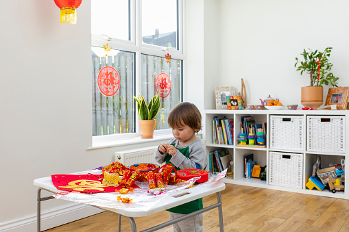 A young boy opening chocolate treats at a table in his living room, the table is covered in decorations for Chinese New Year, ready to decorate the home in Newcastle Upon Tyne, England, patterned imitation fire crackers used to scare away evil spirits are laid out and ready to be hung up.
